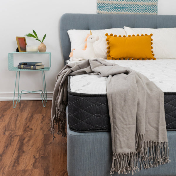 Serta Perfect Sleeper® Reedley Firm Mattress Closeup Corner Shot With Gray Blanket, Unicorn Plushie, And Decorative Pillows. On Bed Frame In Bedroom.