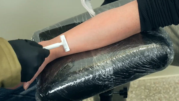 A tattoo artist in gloves is using a disposable shaver to shave a client's arm.