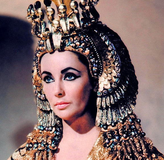 Image shows Elizabeth Taylor from the set of the film Cleopatra wearing a stylish headdress meant to express the sort of ornate emerald-studded jewelry that the historic Cleopatra wore