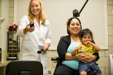 Dr. Crumbliss standing next to a woman and her child who is sitting on her lap in a vision exam chair.