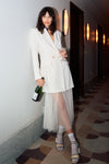 woman leaning on wall holding champagne bottle and glass in white blazer and sheer dress