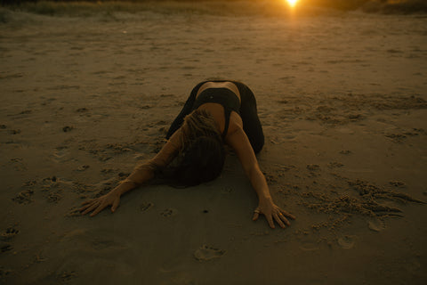 Image of woman kneeling stretched forward on the sand at sunrise.