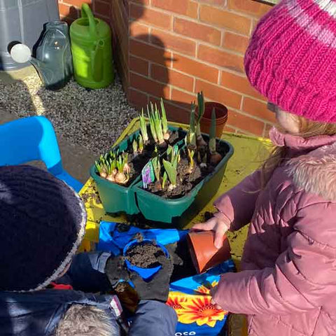 littler girl in pink coat and hat with little boy in blue coat and hat planting bulbs in planters in childrens gardening area