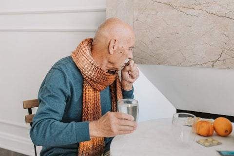 An older man coughing into one hand with a glass of water in his other hand