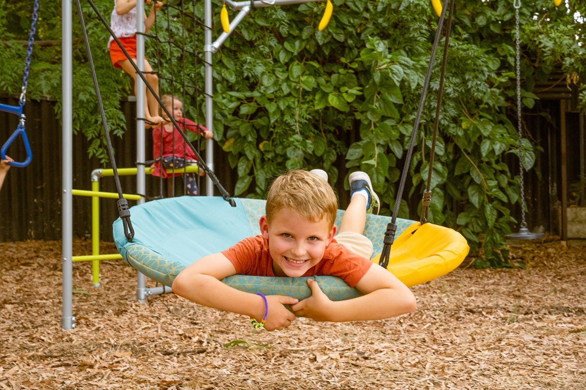 A smiling boy lies on a round nest swing, holding the edge as he enjoys the ride in a leafy backyard. Behind him, kids climb, swing, and play on a Funky Monkey Bars frame over soft mulch in a fun, natural setting.
