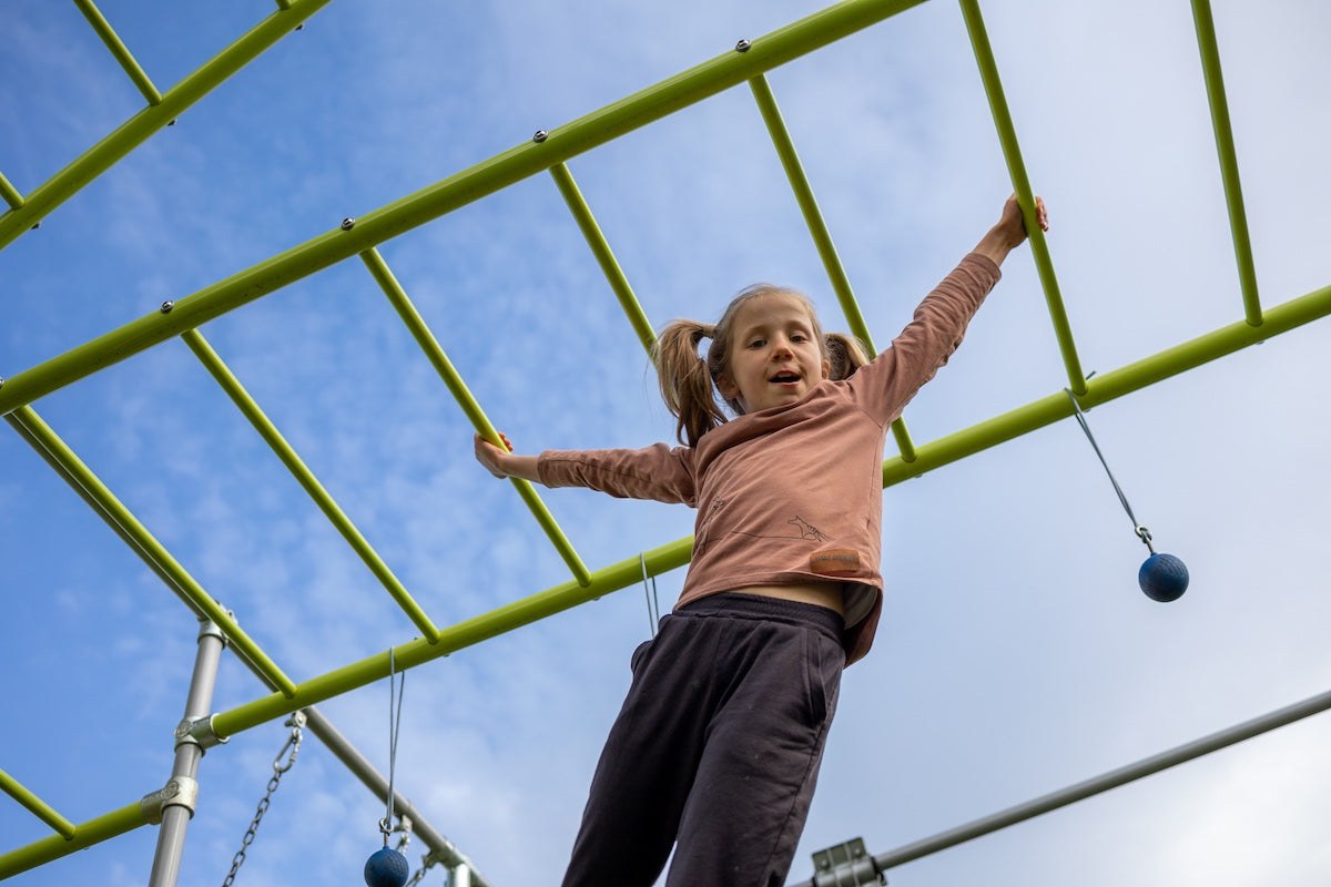 Young girl in brown top swinging across green monkey bars on a Funky Monkey Bars set, smiling with a blue sky backdrop and grip balls nearby.