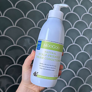 woman holding bottle of MooGoo Cream Conditioner in hand against tiled bathroom wall