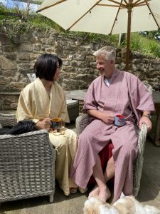 A couple sat outside in the garden on garden furniture. They are both wearing vintage kimono. She is wearing a yellow one and he is wearing a red one.