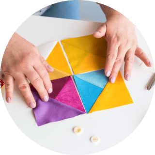 Close-up shot of a person's hands arranging six colorful triangular fabric pieces (yellow, blue, purple, magenta) on a white table to form a hexagon, likely as part of a quilting or patchwork project. Two small buttons are visible nearby.