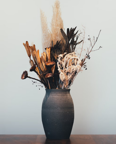 A short, round vase filled with dark blooms like black roses and burgundy dahlias with some greenery mixed in for texture.