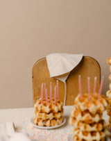 Stacks of waffles with candles on a table with an oat milk brimmed bonnet hanging over a chair