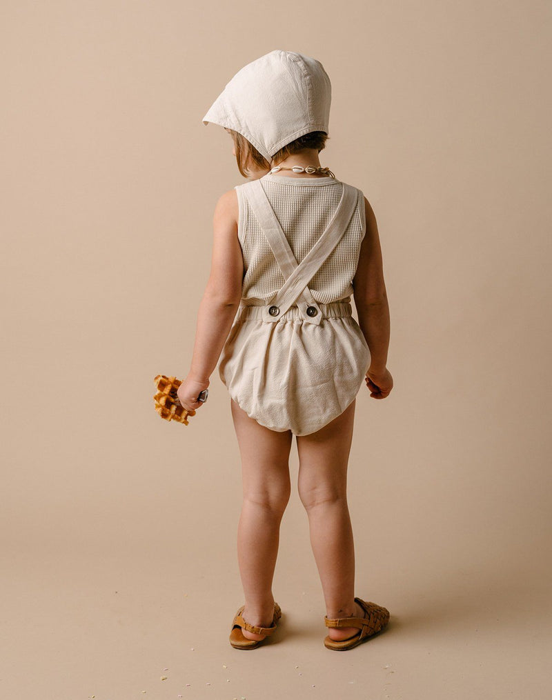 Back of a baby girl wearing an oat milk colored brimmed bonnet, sun suit and waffle tank top