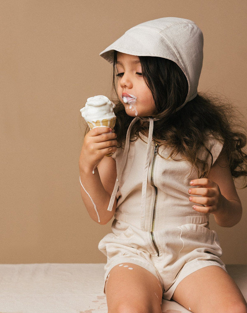 Baby sitting down eating an ice cream cone wearing a bimmed bonnet and tank suit in the oat milk color