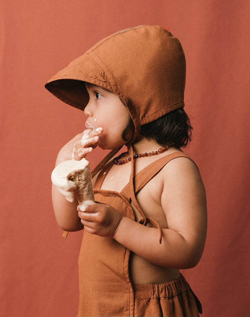 Baby licking ice cream off their fingers while wearing a cinnamon color brimmed bonnet and sun suit