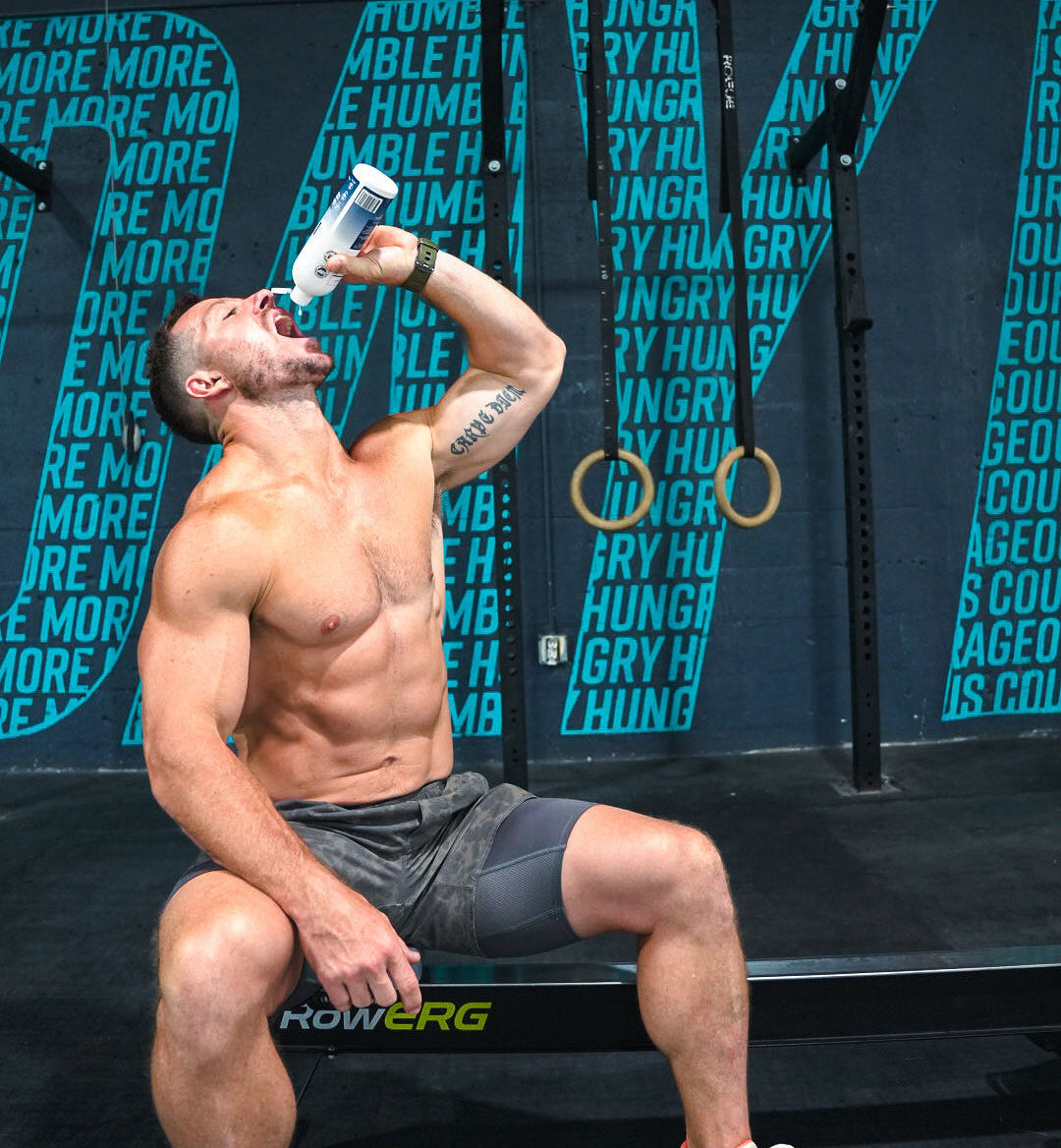 A shirtless man drinks from a bottle while sitting in a gym with motivational text on the wall.