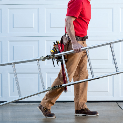 Ein Handwerker in rotem T-Shirt und Khakihosen trägt eine ausziehbare Leiter und hat einen Werkzeuggürtel um. Er scheint sich auf den Weg zu einer Arbeit zu machen.