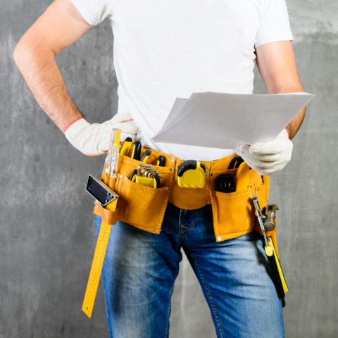 Ein Handwerker mit einem weißen T-Shirt und Jeans steht mit verschränkten Armen und hält in der Hand Pläne oder Papiere, während er einen Werkzeuggürtel mit verschiedenen Werkzeugen trägt.