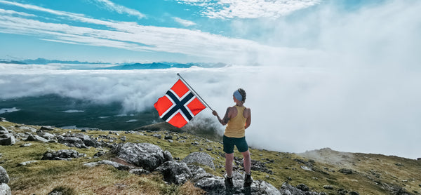 Kvinne på toppen av et fjell med lavt skydekke. Hun holder et svaiende norsk flagg og speider utover mot det hvite skydekket som ligger foran henne med blå himmel rundt seg.