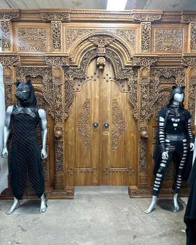 Two mannequins wearing a leather outfit and a vegan leather outside in front of an ornately carved wooden door from Bali.