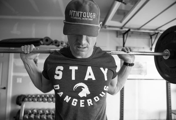 A man wearing a MTNTOUGH shirt and hat, loading up for a squat underneath a barbell