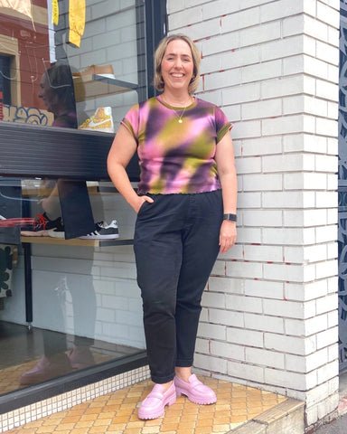 Emma standing in front of Mutual Muse Brunswick, against black floor-to-ceiling windows to the left of the image, and a white brick wall to the right. She poses with her right hand in her pocket and smiles at the camera. She has shoulder-length blonde hair, and wears a t-shirt in gradient shades of olive, pink, and purple, as well as black pants and lilac loafers.