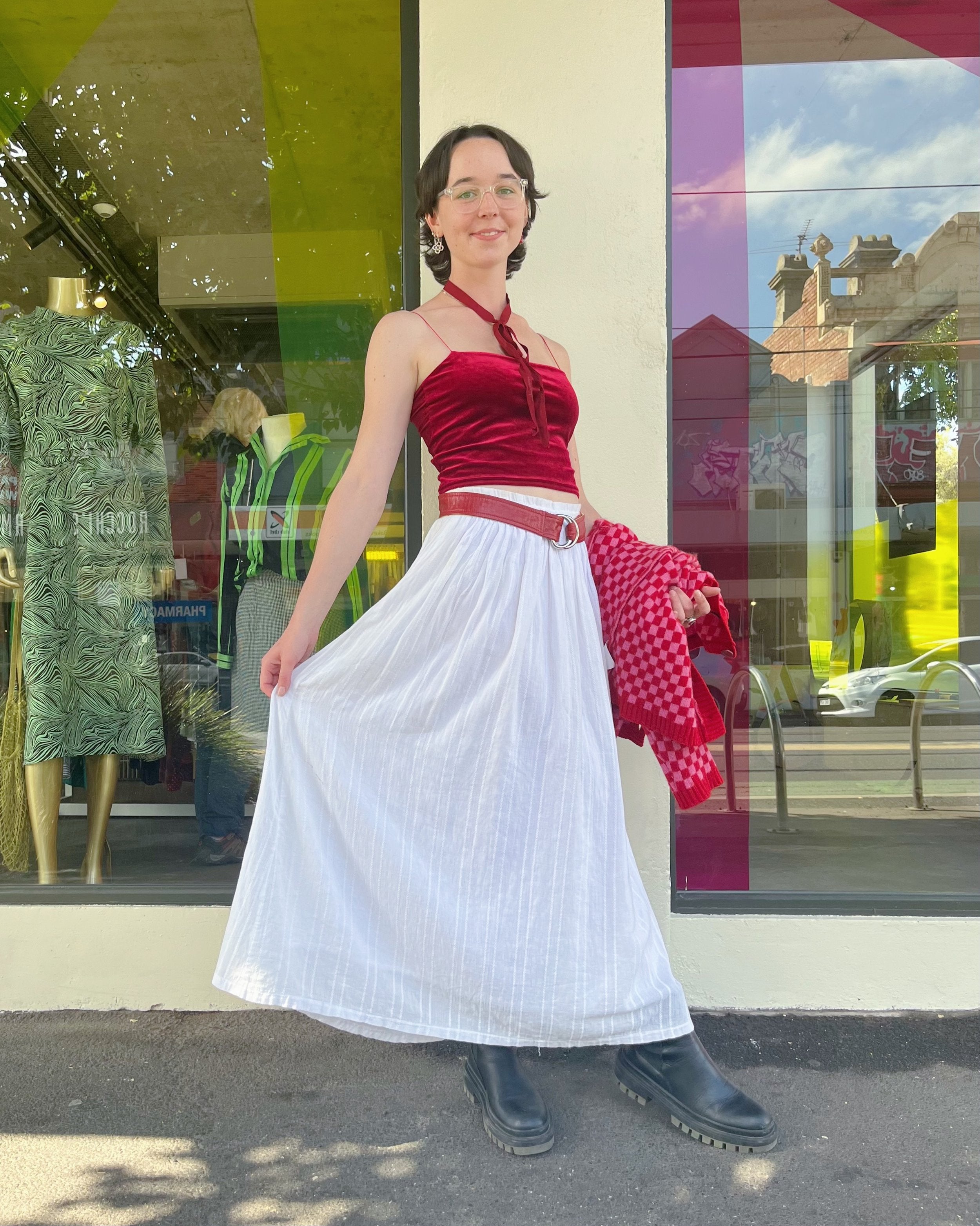 Chessie standing in front of Mutual Muse Northcote, posing side-on and holding out her skirt with one hand. She is wearing a dark-red velvet tank with a matching red ribbon around her neck. She also wears a red belt, a white skirt, and black boots. In her left hand, she holds a red-and-pink checkered cardigan. Behind her is the Mutual Muse Northcote storefront, with floor-to-ceiling windows and white walls.