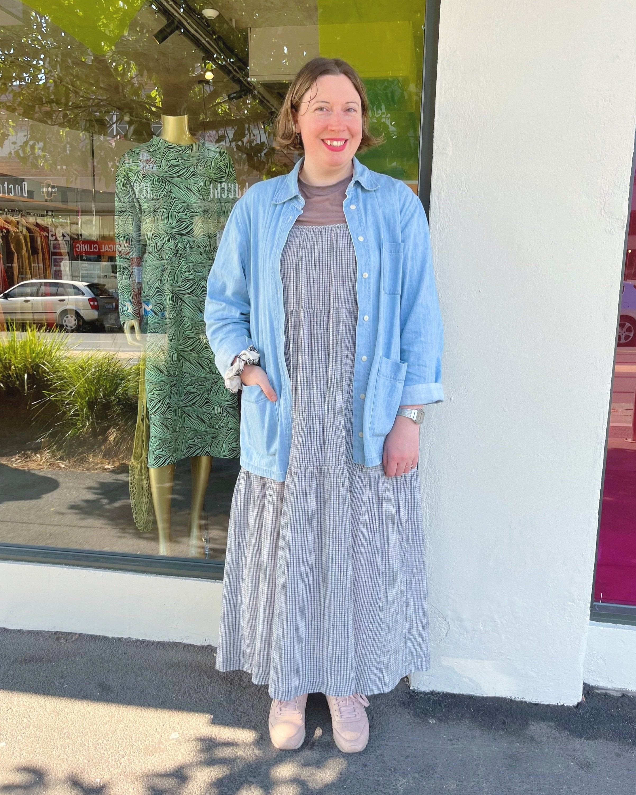 Sarah stands in front of Mutual Muse Northcote. In the background is the store’s floor-to-ceiling windows and a white pillar. Behind the window, we can see the clothing in store and reflections of the street. Sarah wears a brown t-shirt, a long checkered dress, a lightwash denim jacket, and lilac sneakers. Her face is slightly tilted to her left. She smiles at the camera.