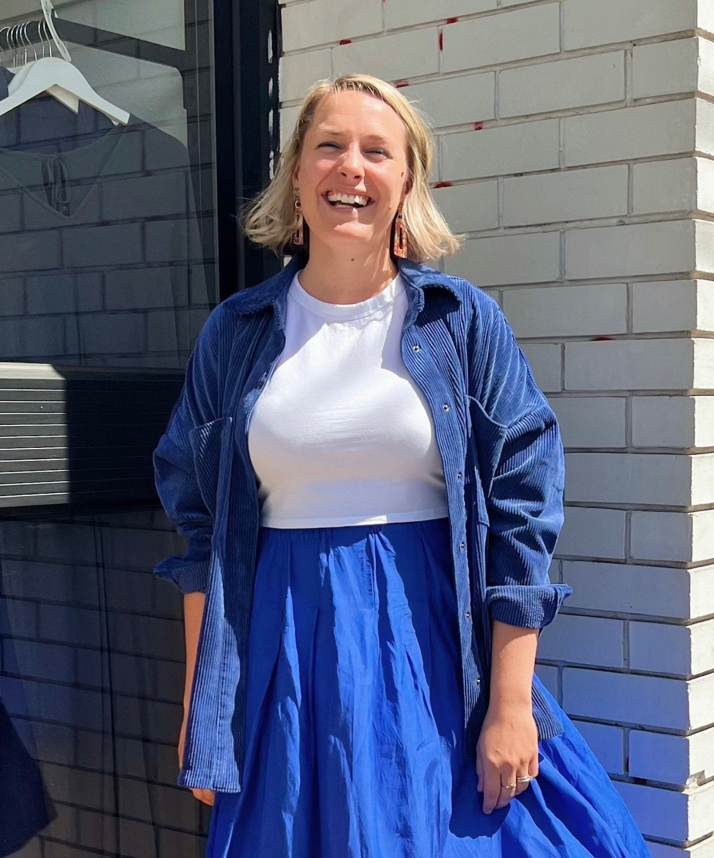 Sara standing against the sunlight in front of MM Brunswick’s white brick storefront. She has golden shoulder-length hair and smiles at the camera. She is wearing a white top, blue corduroy button-down shirt, and blue skirt.