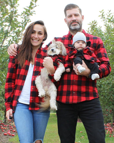 Mom, dad, baby, and small dog standing outside in front of some trees all wearing matching red and black buffalo plaid shirts