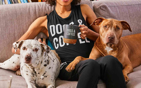 woman holding mug that says "dogs and coffee" with a mid-size mixed breed dog sitting on each side of her on a sofa