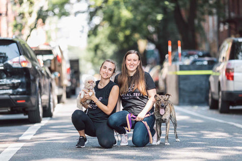 Two women in Social Tees t-shirts with two dogs crouching in a New York city street