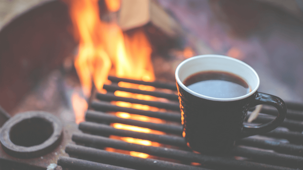 Coffee mug on campfire grill, flames in background, cozy outdoor camping scene