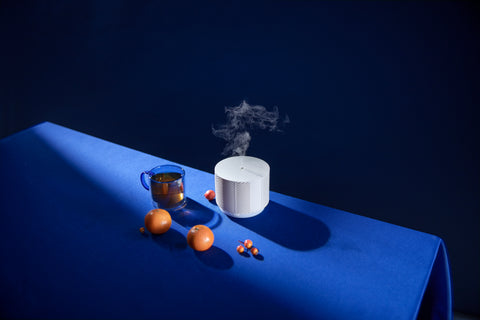 An aroma nook essential oil diffuser placed on a blue tablecloth next to a cup of tea and two clementines