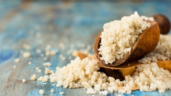  The image displays a scoop full of coarse, granular white salt, commonly referred to as Celtic sea salt, resting on a wooden surface with a blue-hued background. The salt crystals appear irregular in shape and size
