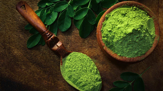 The image showcases Moringa oleifera in two forms: fresh, vibrant green leaves and as a finely milled powder, likely intended for consumption. The powder is contained in a rustic, earth-toned bowl with a wooden spoon, suggesting its organic quality. 