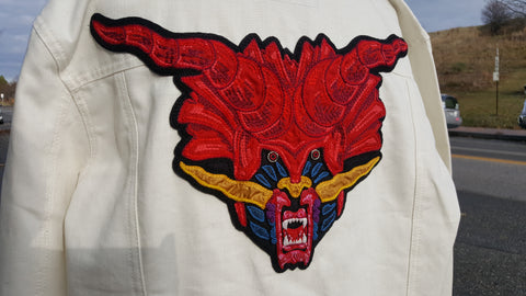 Chainstitch and applique back patch of the creature's face from the Defenders of the Faith album cover on a white denim jacket. It's large red horns extend from shoulder to shoulder, with it's lower jaw landing at the center of the back. It's eye sockets and cheeks are a royal blue, with a gold nose and whiskers. It's snarling lips and chin texture is created with red and pink embroidered knots and it has sharp white teeth.