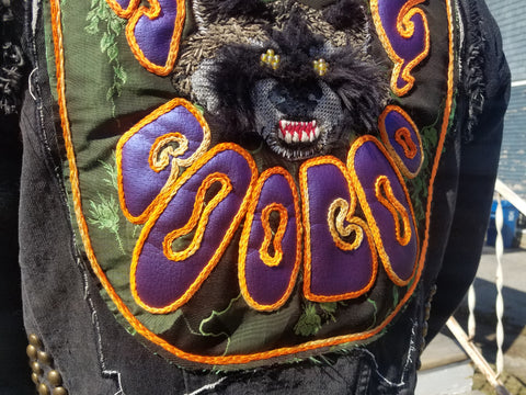 A closeup of the text at the bottom of the back patch, reading "Boo Boo". The text is a thick, 70's style 'groovy' font, filled with a metallic purple vinyl and outlined with a variegated orange chainstitch. 