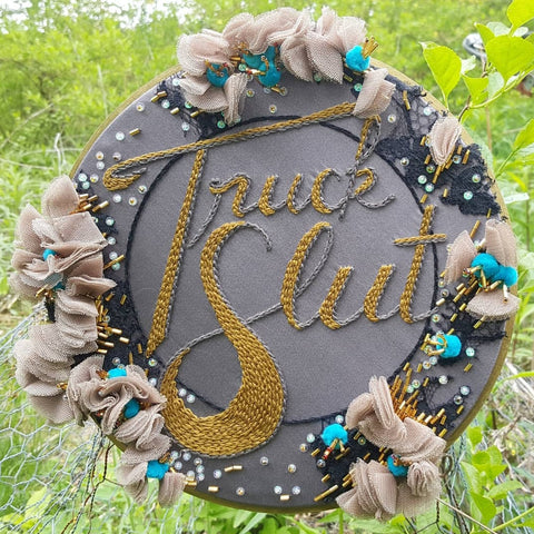 A decorative embroidery hoop. The base fabric is a dusty lavender satin, the wide border is collaged with black lace, blue pom poms, beige folded fabric, opalescent and gold beads and iridescent sequin. At the center of the hoop is an olive green chainstitch reading "Truck Slut", the script is outlined with a grey thread.  