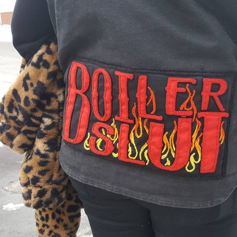 A rectangular tramp stamp patch that reads "Boiler Slut". The background of the patch is black and the lettering is made up of a red applique, outlined with red chainstitch. Behind the word "Slut" is a chainstitched outline of 90's style graphic flames, that fade from red at the top of the flames down to orange and yellow at the base. 