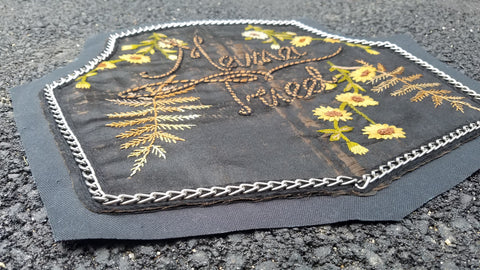 A foreshortened image of the bottom left corner of a back patch, laying on the pavement. The patch is black with a thin metal chain border, "Mama Tried" is embroidered at the center of the piece in brown thread, outlined with a black twisted rope detail. The base fabric is a salvaged black silk that already had hand embroidered daisies and ferns.