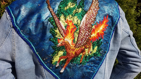 A large blue, upside down triangular shaped patch on a denim jacket. The patch stretches across the shoulders of the jacket, with the third point of the triangle landing just below the wearer's rib cage. The hand stitched embroidery is of a pegasus in flight with a flaming mane and tail, in front of a beaded applique bush. It's extended wings are made up of ivory, burgundy, olive and lavender hues, while the pegasus itself is a mix of warm browns and a couple white accents. There's a smattering of rhinestone studs throughout the blue background.