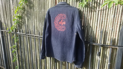 Backside of a dark blue denim work shirt, centered between the shoulders is a logo in red chainstitch. The design is of a chainlink wreath design, with an opening in the links at the right side of the circle, allowing lowercase red text reading "cbja" to break through. 