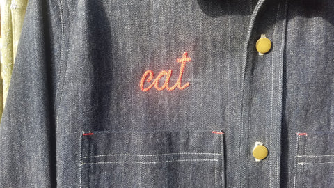 Red, chainstitch scrips reading "cat". Placed on a dark blue denim work shirt, just over the pocket.