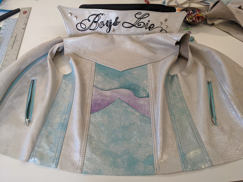 A collaged leather jacket made by artist Gnykol. On the backside of the collar is black chainstitch script reading "Boys Lie", that is exposed when the collar is flipped up.
