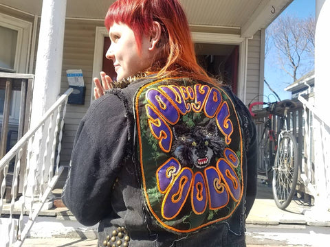 A full view of a chainstitch and applique back patch on a studded denim vest, worn by a non-binary person with a red mullet. The patch reads "You Do You Boo Boo", the text wraps around the top and bottom of the patch, framing a snarling raccoon face at the center.