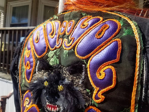 The top third of a chainstitch and applique back patch, sewn onto a studded denim vest. The text along the top of the patch reads "You Do You", it's a metallic purple vinyl applique, outlined with variegated orange chainstitch. The background of the patch is a green and black satin, with a mixed media snarling raccoon face at the center.
