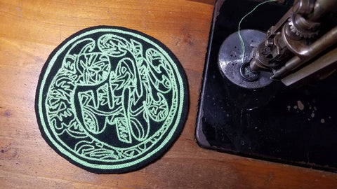 A circular chainstitch patch resting on the worktable next to the machine head. The patch has a black background with neon green thread, the design is of a swirling font reading "Cunt". The lettering is filled in with an abstracted line work floral design.