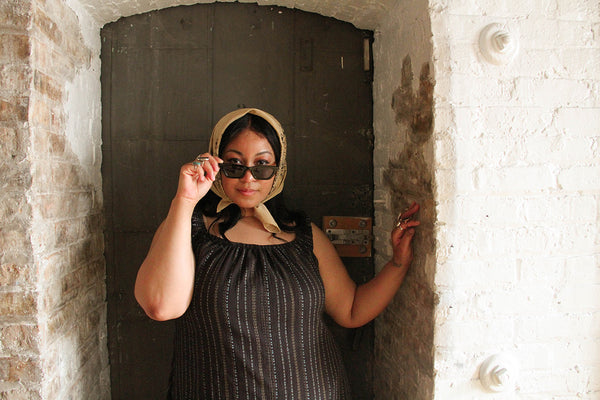 A plus size model wearing a prairie dress and silk headscarf looks at the camera over her sunglasses