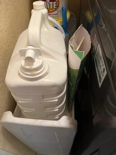 Large white detergent bottle and cardboard box beside a washing machine in a laundry area.