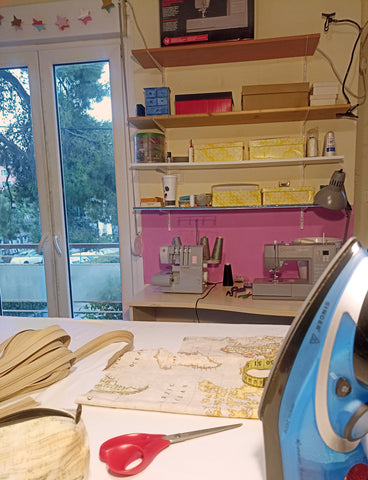 Eirianna's current sewing workshop. There is a large table with fabrics and an iron on it. There is also a sewing machine and a serger on  a desk. Above the desk there are shelves with boxes on them.